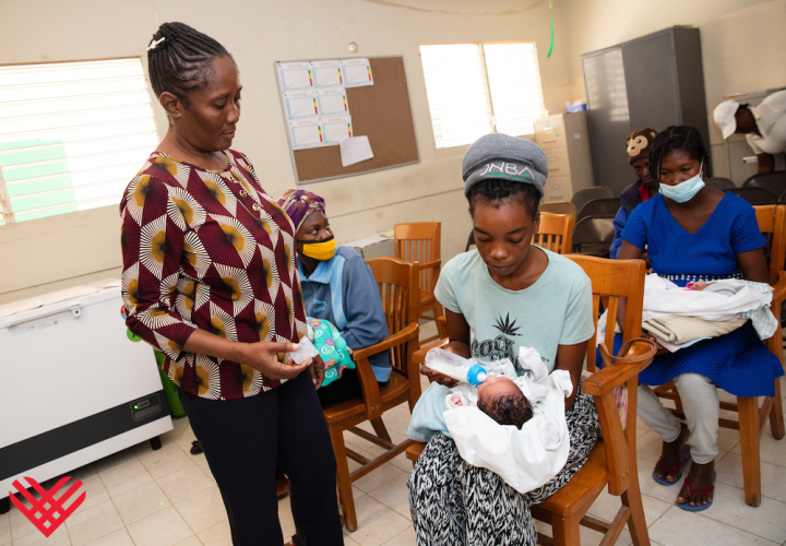 A Haitian clinician wearing a maroon, white, and black patterned shirt talks with a young mother who is sitting in a chair cradling her baby. 