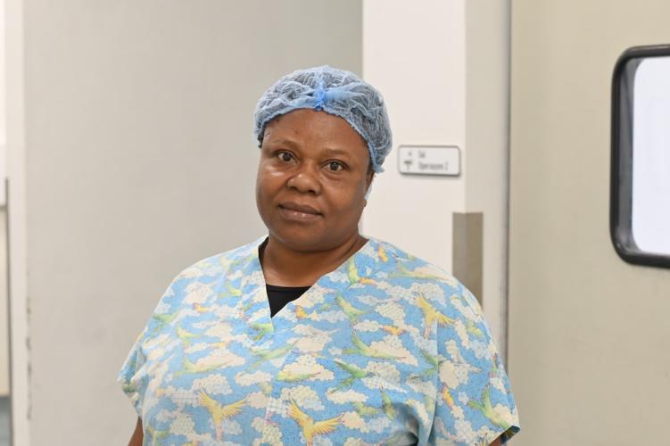 A woman Haitian nurse anethesist stands in front of a doorway wearing light blue scrubs and a blue hairnet. She smiles at the camear.