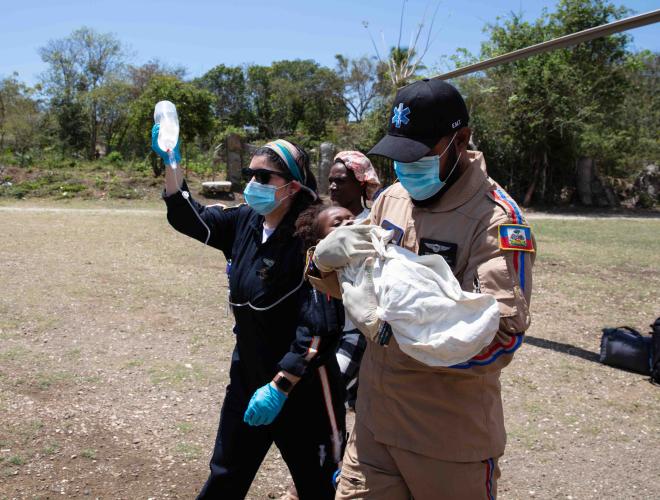 A uniformed male EMT carries a baby swaddled in white across a dry dirt field. Beside him walk two women, one holding up an IV bag.