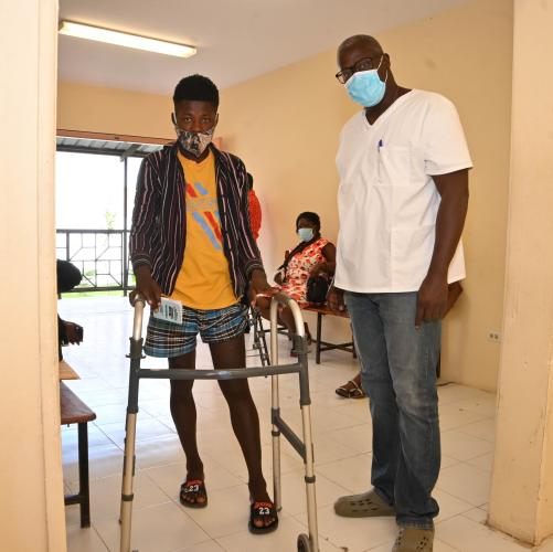 A young man stands, holding a walker, next to an older, taller man wearing glasses and a white scrub shirt. Both wear face masks.