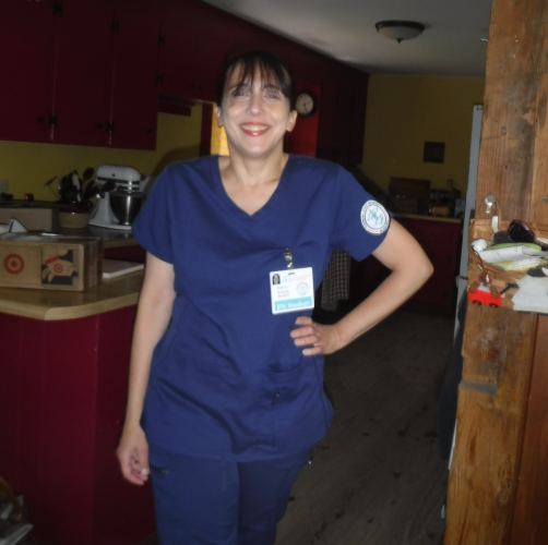 A light skinned woman stands in a wood-framed doorway dressed in dark blue medical scrubs. She has one hand on her hip and smiles at the camera.