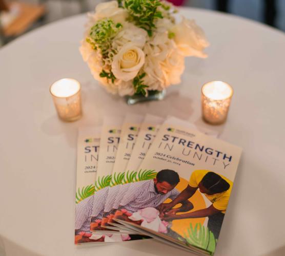 A round tabletop holding a bouquet of white flowers, 2 candles, and a stack of programs with the words “Strength in Unity” and a cutout photo of an infant being vaccinated.