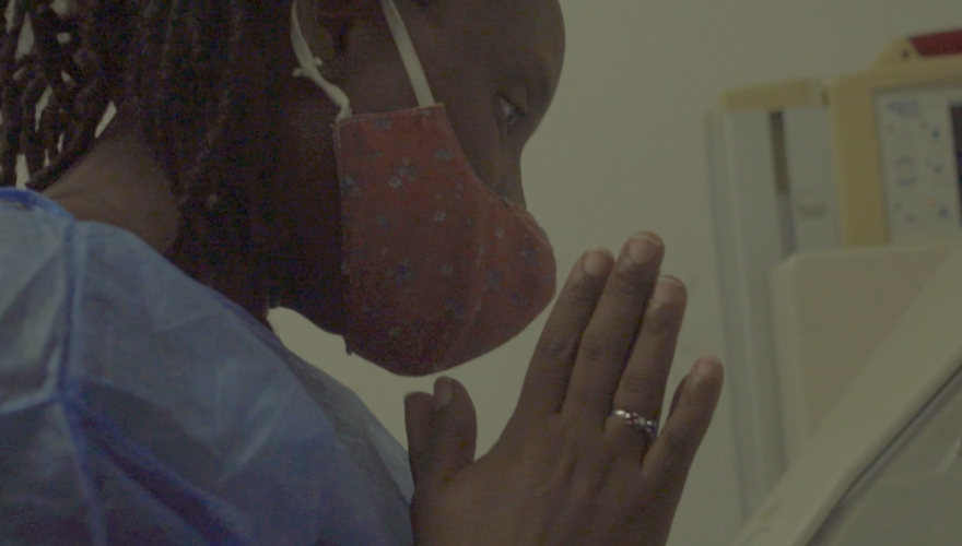 Close profile of a Haitian woman gazing downward, hands in prayer. She has braids and wears a flowered face mask, blue hospital gown, and silver ring.