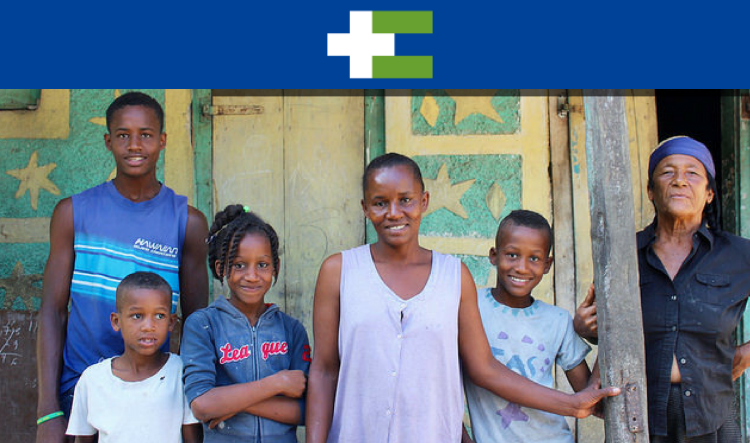 A six-member Haitian family stands in front of a house. The mother stands in the middle with three young children by her sides. An older woman stands to the right and a tall man stands to the left.