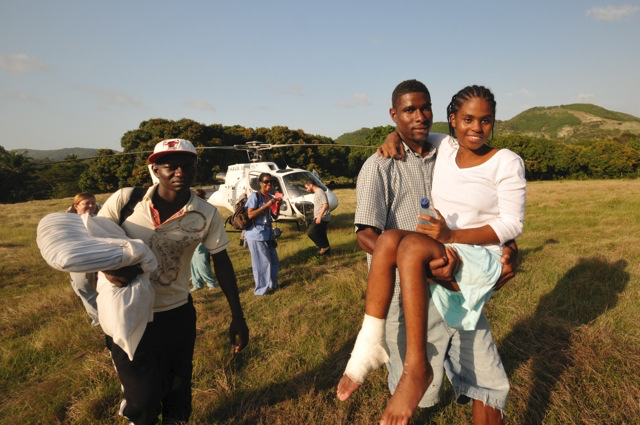 A Haitian man carries a woman across a field. The woman has his arm around his neck and has a bandage on her foot. A helicopter stands in the background.