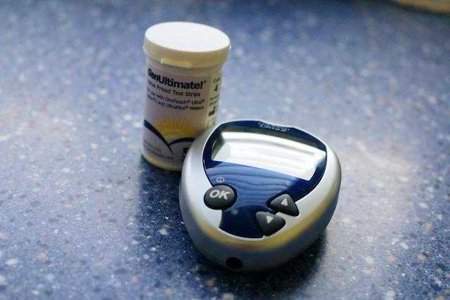 A small handheld diabetes glucometer and a cylindical container of testing strips sits on a speckled blue countertop.