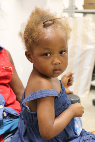 A young female Haitian child sits in sideways and turns her head to face the camera. She has short, light-colored hair and wears a light blue dress.