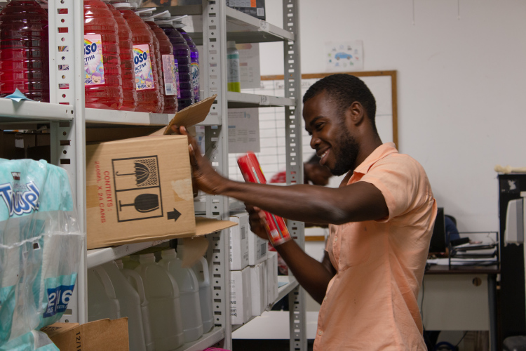 A Haitian man stands in front of a metal shelving unit. He reaches into a cardboard box to put away a canister.