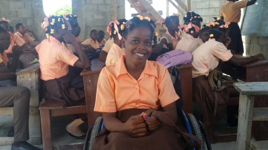 A girl in a peach-colored shirt and hair ties smiles happily at the camera. She sits in a wheelchair in a cinderblock classroom with other students.