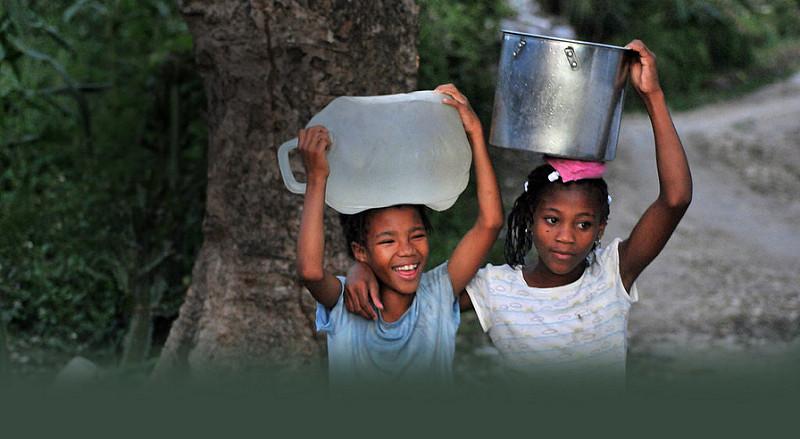 two young girls carrying bucks of water on their heads and smiling