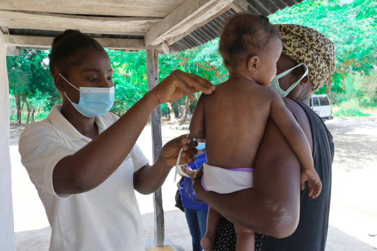 Three people appear in the picture. On the left, a community health worker wears a yellow polo shirt and wears a blue medical mask covering her mouth and nose. She holds a malnutrition screening strip against the arm of a young child, who is in the center of the photo. The baby wears a white diaper. The baby is held by an adult woman on the right side of the photo. The woman wears an animal printed head wrap and a black dress.