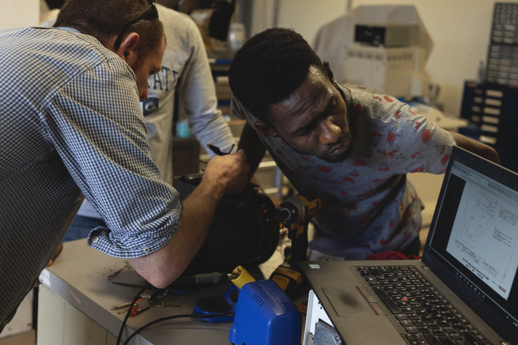 Two men work on repairing a motor. A light-skinned man adjusts something while a dark-skinned man leans in to listen.
