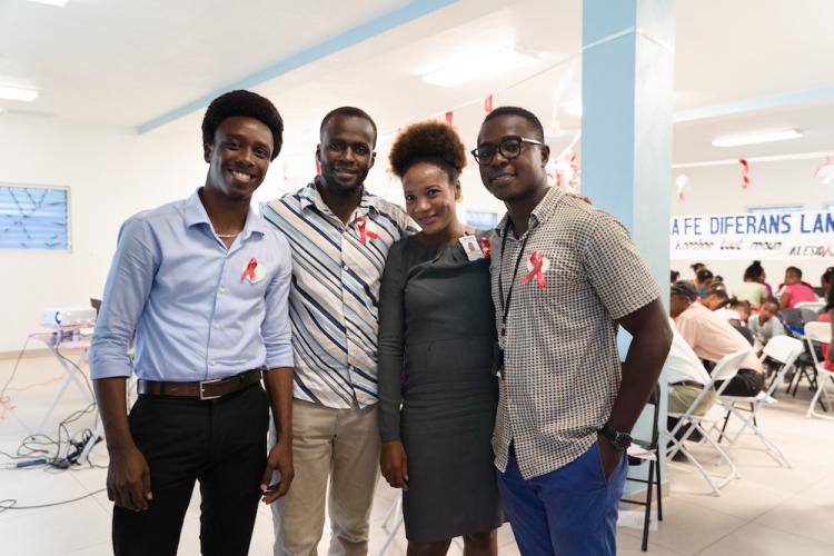 Four Haitians in business casual clothes stand together in a large, bright room, smiling and looking relaxed.