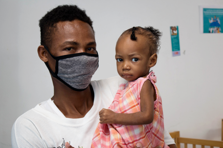 A Haitian man wearing a grey cloth face mask holds his baby daughter in his arms. She is wearing a pink plaid dress and has her hair up.