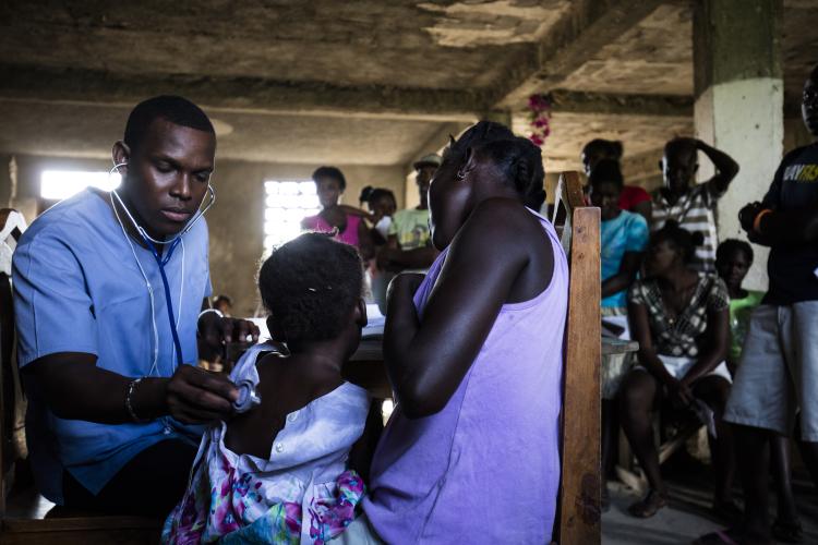 A male Haitian doctor examines two patients at a crowded community health clinic.