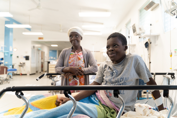 A young Haitian boy props himself up in a hospital bed. He smiles at the camera. His mother stands behind him with her hands together, also smiling.