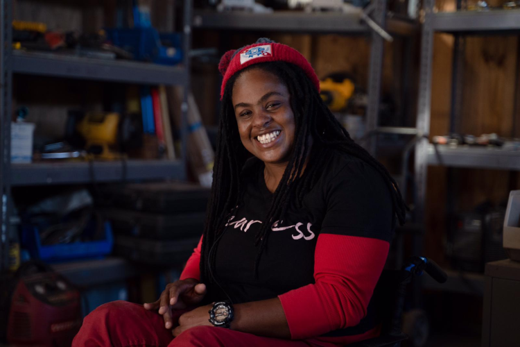 A Haitian woman sits in a wheelchair in an equipment workshop. She smiles brightly at the camera.