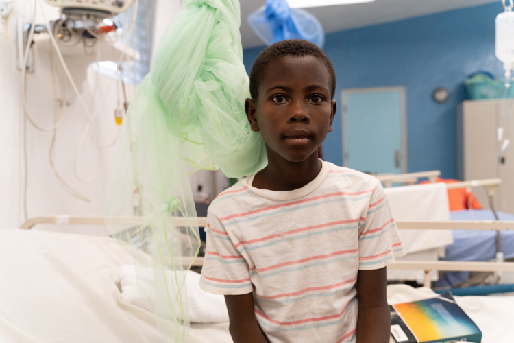 A young Haitian boy wearing a striped t-shirt sits on a hospital bed. A light green mosquito net is tied up behind him.
