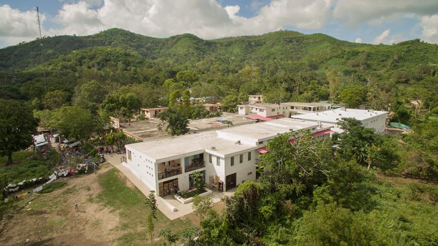 Aerial view of hospital buildings in rural Haiti. The buildings are mostly two storey and are nestled in a valley of green trees.