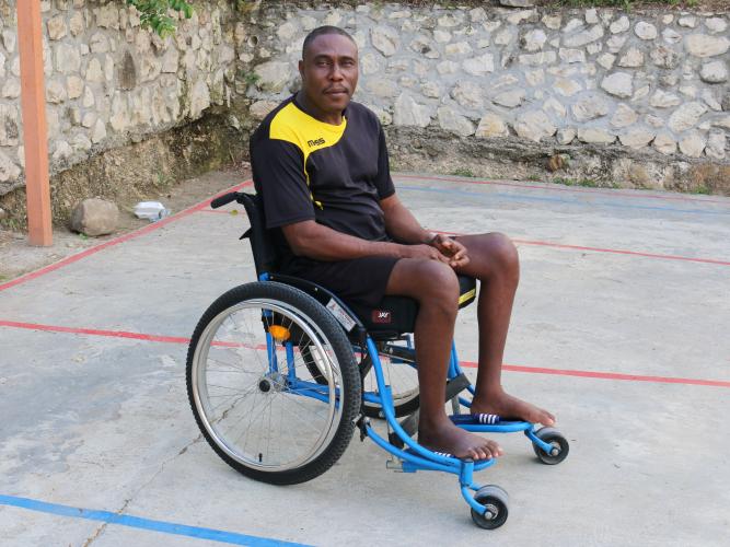 A Haitian man sits in a wheelchair on a concrete basketball court. He turns his head to the side to look at the camera.