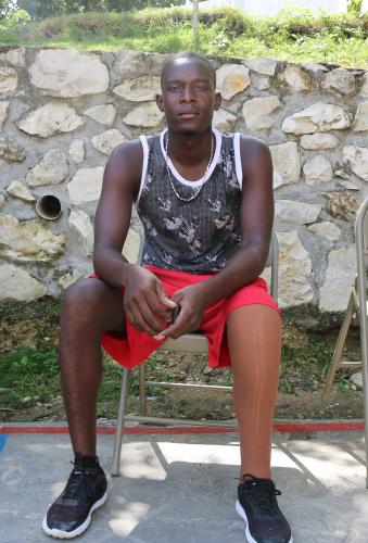 A tall Haitian man sits in a metal folding chair looks at the camera with his hands in his lap. His left leg is a prosthetic.