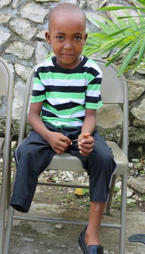A young boy who is missing his right leg sits in a metal folding chair. He wears a blue, white, and green striped shirt and short black pants.