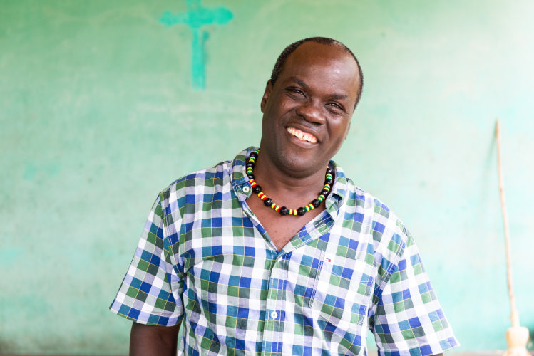 A Haitian man wearing a beaded necklace and a checkered shirt stands in front of a green wall and smiles.