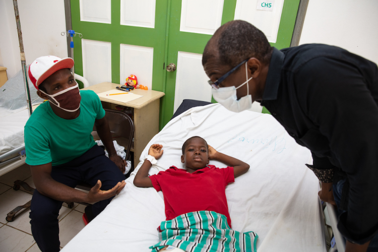 A young boy lays in a hospital bed. His father sits in a chair to the left of the bed and a doctor stands to a right. The doctor and the patient's father are talking.