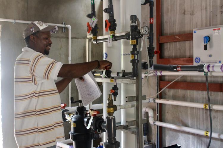 A man stands to the left of a series of thin pipes that are horizontal to a grey cement wall. He adjusts one of many valves on the pipe system.