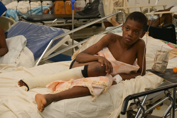 A young boy lies in a hospital bed with a leg injury. He is covered by an orange and white blanket. His right leg is wrapped in a bandage below the knee.