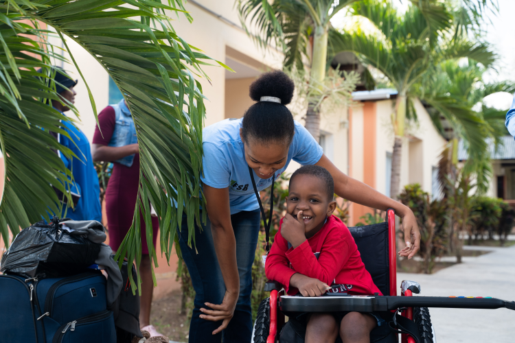 A young Haitian boy sits in a wheelchair and leans to the left to whisper to a female clinician, who stands next to him.