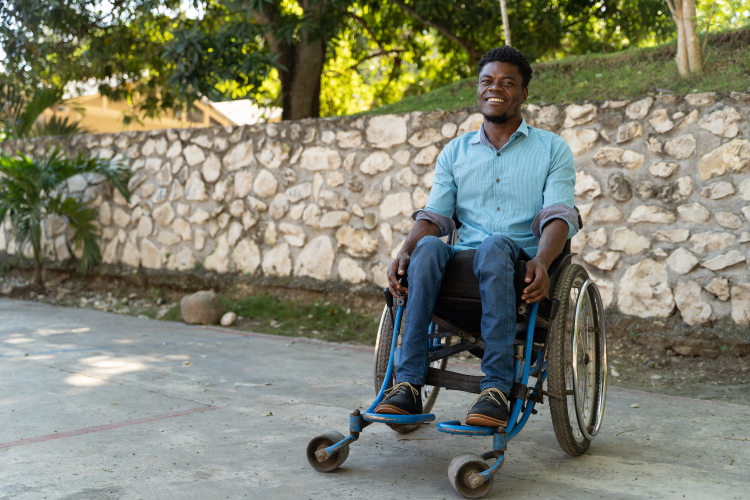A Haitian man in a wheelchair sits in front of a wall, behind which are lush green trees. The man smiles brightly.
