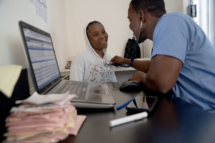A Haitian doctor wearing blue scrubs sits at a desk near an open laptop computer. A female patient sits in a chair besides him and smiles. He holds a stethescope to her chest.