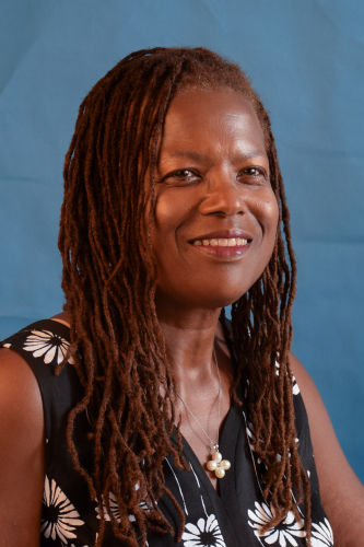A headshot of Dr. Michele David smiling. She has locs and wears a floral blouse and is situated against a blue background.