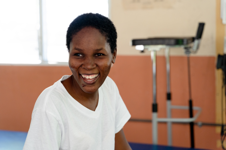 A Haitian woman smiles at the camera. She is shown from the waist up in a physical therapy office.