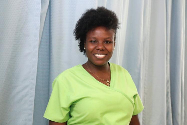 A female Haitian clinician stands in front of a blue curtain. She is wearing lime green scrubs. She smiles at the camera.