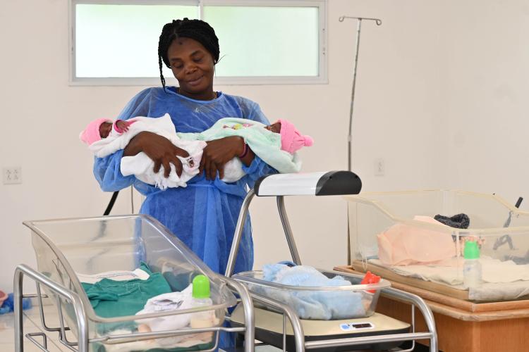 In a hospital room, a Haitian woman in a blue surgical gown holds two newborns with pink hats. A third newborn rests in a bassinet. 