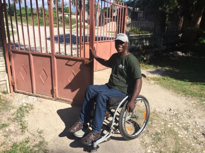 A Haitian man holds open a peach-colored metal gate outside on a dusty road.