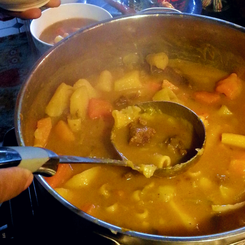 A pot of soup joumou, Haitian pumpkin soup. The soup has a thick broth, with potatoes, yams, and beef. A ladle dips into the pot.