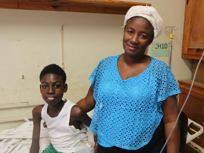 A young Haitian boy sits on a hospital bed and smiles. His mother stands next to him, with her arm around his shoulder.