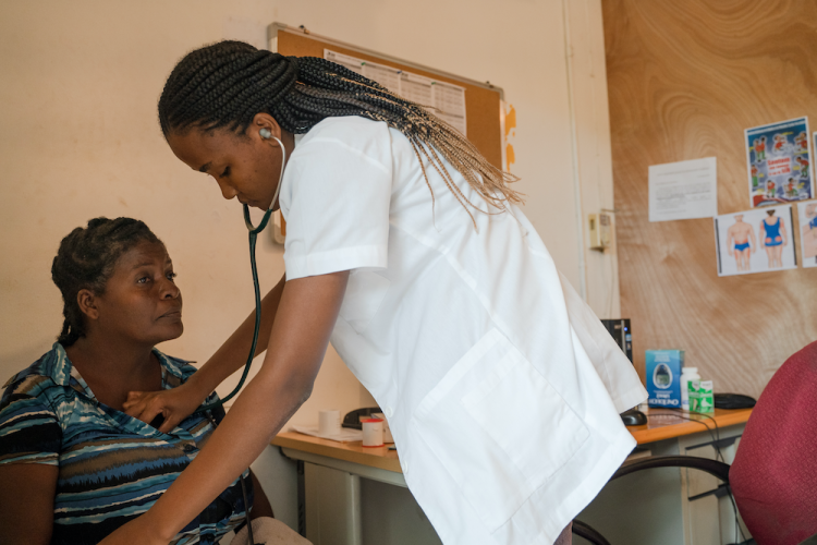 A female Haitian doctor wearing a white coat stands in front of a female patient. She leans over to listen to the patient's chest with a stethescope.