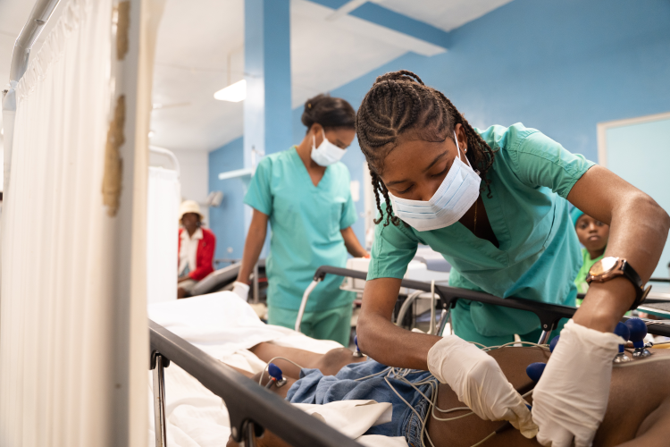 A young female Haitian clinician wearing a medical mask bends over a patient in a hospital bed, adjusting some sensors on the patient's side.