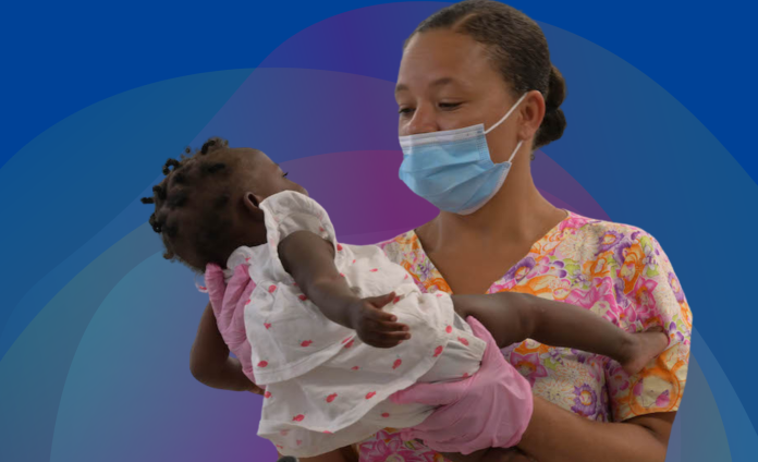 A Haitian nurse wearing a blue medical mask, a flower-printed scrubs top, and pink medical gloves holds up a girl Haitian baby, who has short hair and wears a white dress with pink flowers.