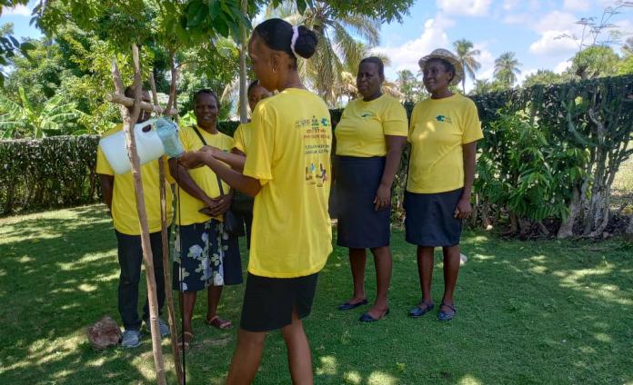A woman wearing a bright yellow t-shirt demonstrates how to use a handwashing station. A group of five women in the same yellow t-shirt stand around her and observe.