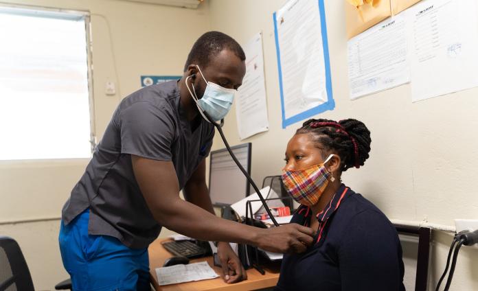 A Haitian doctor stands checks a female patient's breathing with a stethescope on her chest. The patient sits in a chair next to a desk.