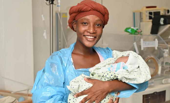 A young mother wearing a dark orange head wrap and a blue medical gown cradles her baby in the NICU.