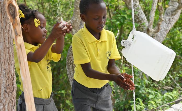 A young boy and girl in yellow school polo shirts wash their hands using a Tippy Tap device.