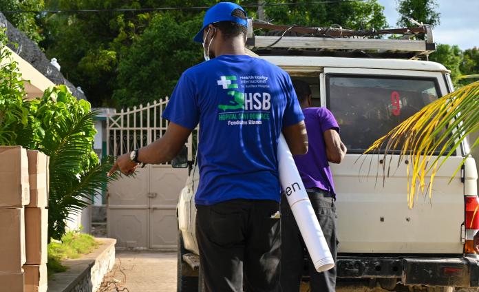 In a courtyard, men unload cardboard boxes from a 4WD vehicle. 1 wears a bright blue cap and T-shirt with HEI/SBH logo.