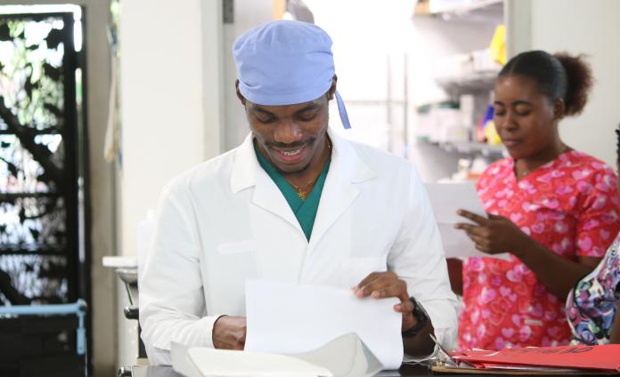 In a bright hospital space a man in a lab coat and a woman in red scrubs with pink hearts look down at papers in their hands, smiling.