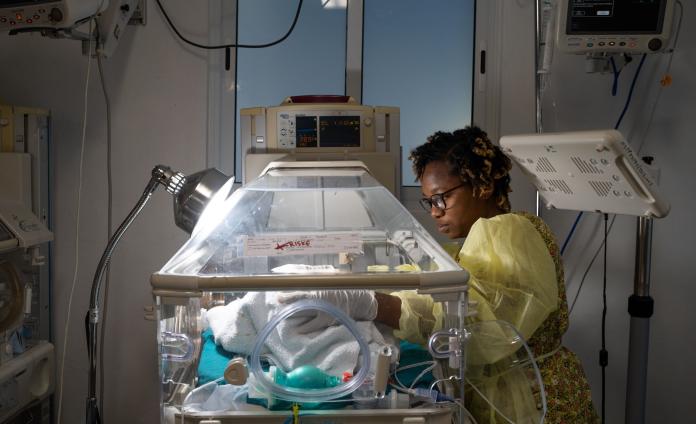 In a darkened hospital room, a nurse focuses her attention on an infant in an incubator lit by a warming light. 
